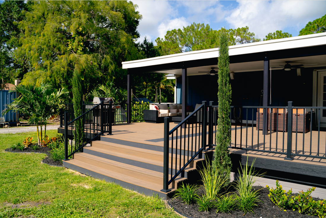A modern home with a spacious covered deck, black railings, outdoor seating, and lush landscaping under a sunny blue sky.