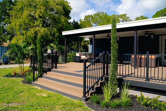 A modern home with a spacious covered deck, black railings, outdoor seating, and lush landscaping under a sunny blue sky.