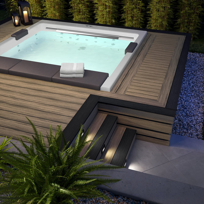 Hot tub with wooden exterior and towels inside, surrounded by plants and lanterns at night.