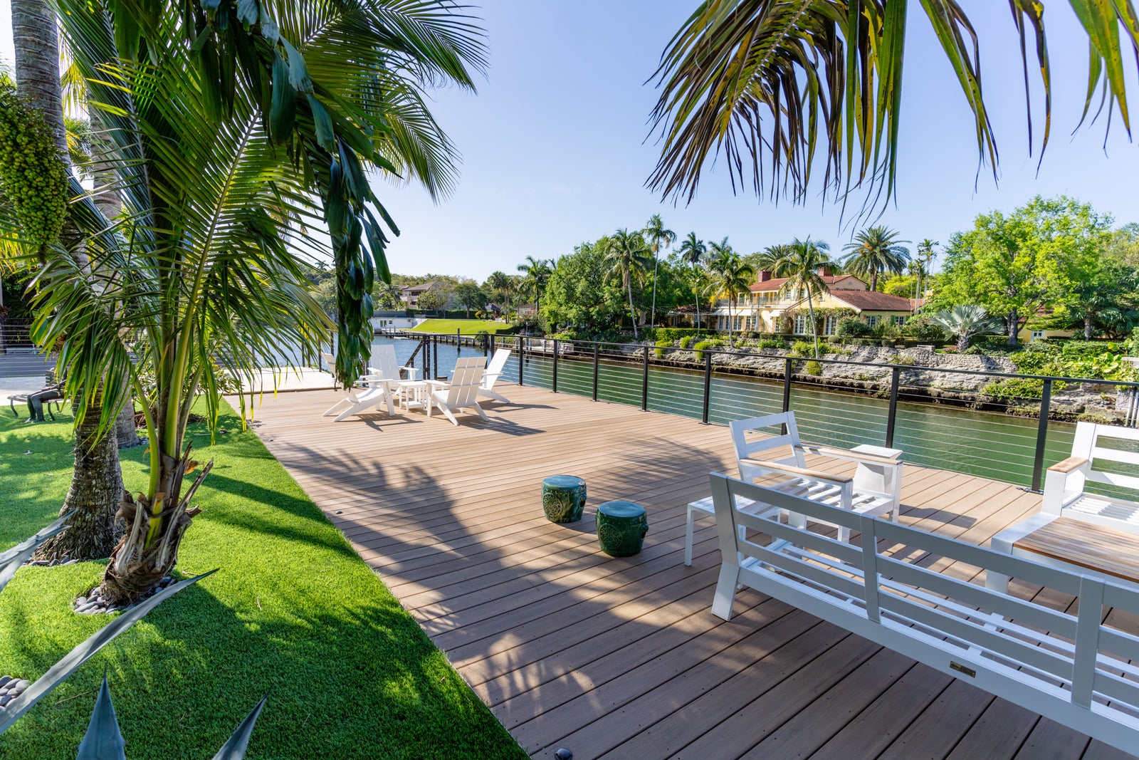 Outdoor deck with furniture by a waterfront, surrounded by greenery and palm trees.