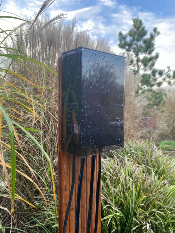 Black rectangular object on a wooden post with grass and trees in the background
