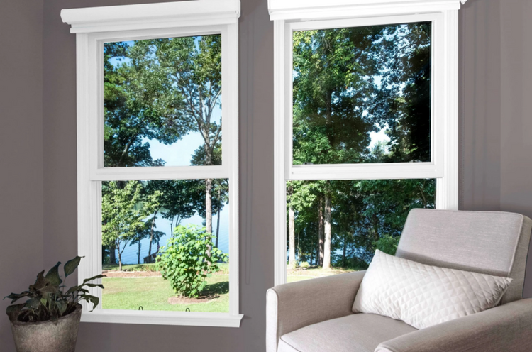 Two white double-hung windows with a view of trees and a gray chair in front.