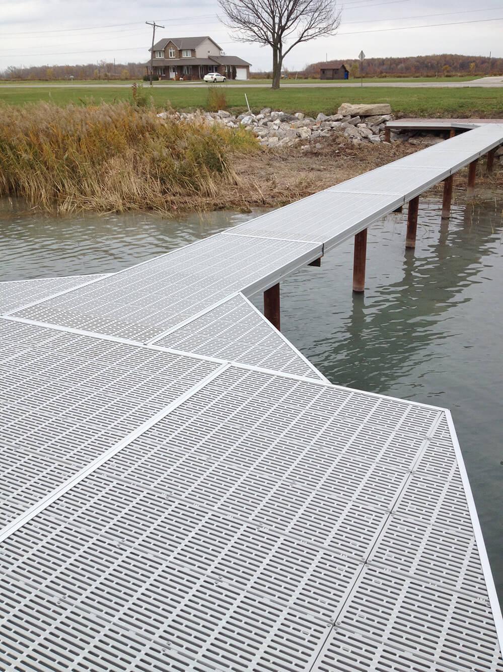 Dock with metal grating extending into a body of water, with a house and trees in the background.
